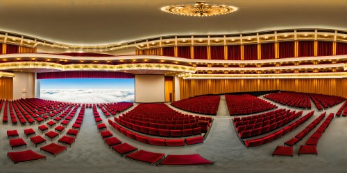 A grand concert hall within the Kennedy Center in Washington, D.C., boasts opulent red velvet seats under a shimmering chandelier, reflecting golden light against intricate, ornate ceilings.